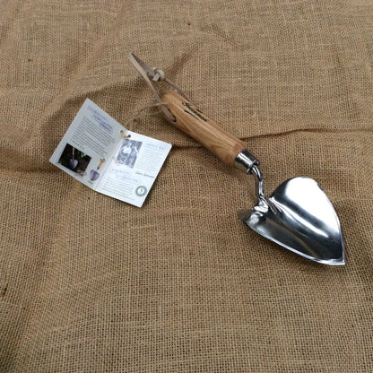 A small hand trowel with a polished silver metal blade and a light brown wooden handle, displayed on a textured burlap fabric background next to its paper instruction booklet.
