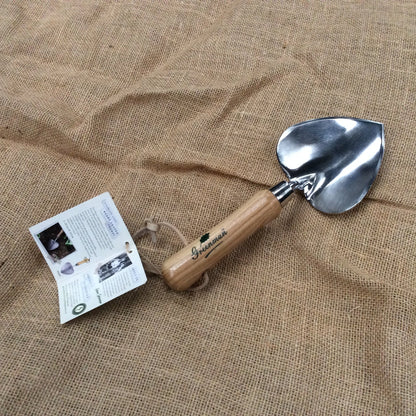 A small hand trowel with a polished steel blade and a light wood handle, resting on a textured burlap fabric background next to its paper tag.