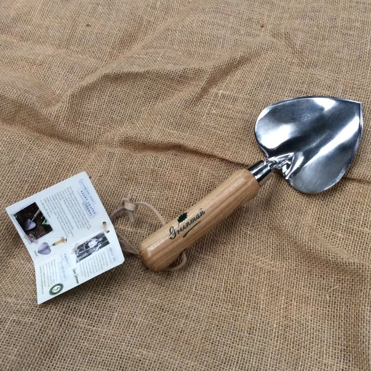 A small hand trowel with a polished steel blade and a light brown wooden handle, resting on a textured burlap fabric background next to its paper tag.