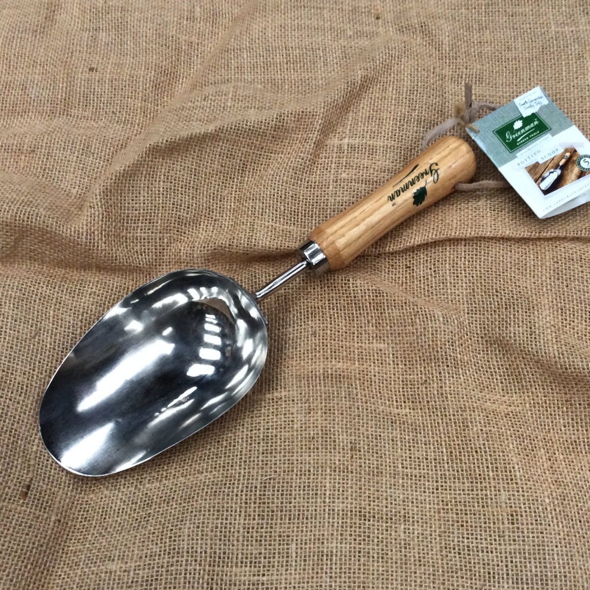 A metal hand trowel with a light wood handle and a small attached product tag, placed on a textured tan burlap fabric background.