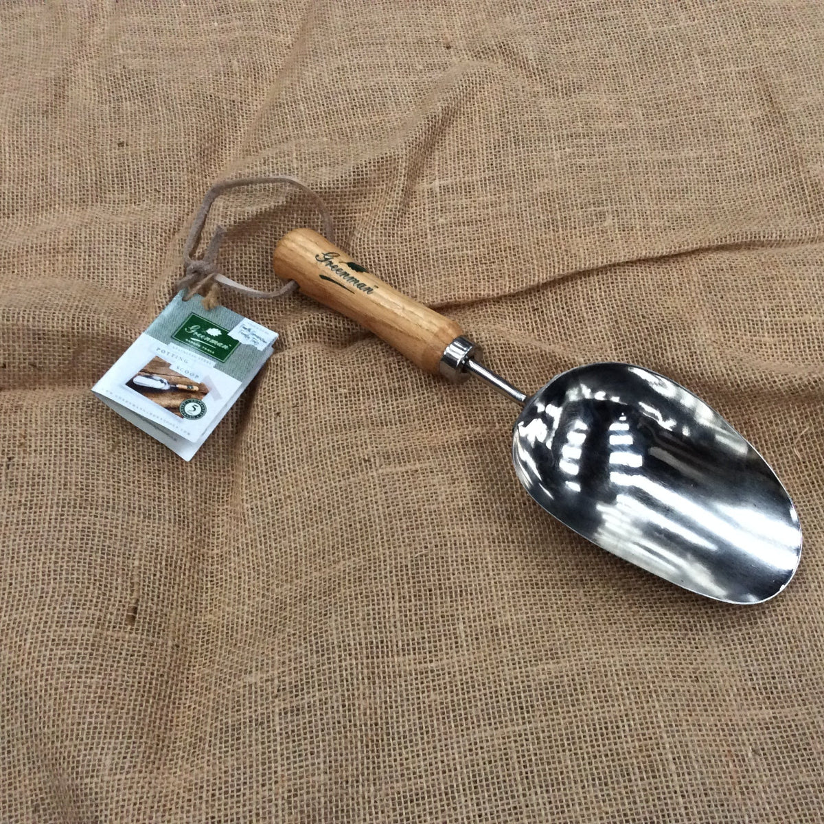 A small hand trowel with a polished silver metal scoop and a light brown wooden handle, resting on a textured brown burlap fabric background. The trowel is attached to a paper hangtag.