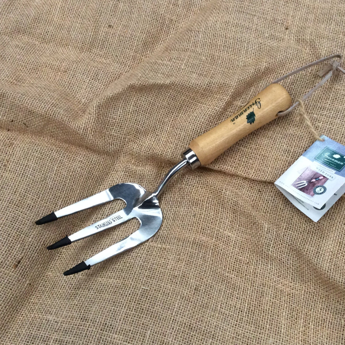 A metal three-tooth hand fork garden tool with a light wood handle, placed on a burlap fabric background next to its product tag.