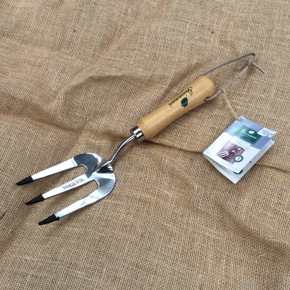 A metal three-prong garden hand fork with a light wood handle, displayed on a textured burlap background next to its product tag.