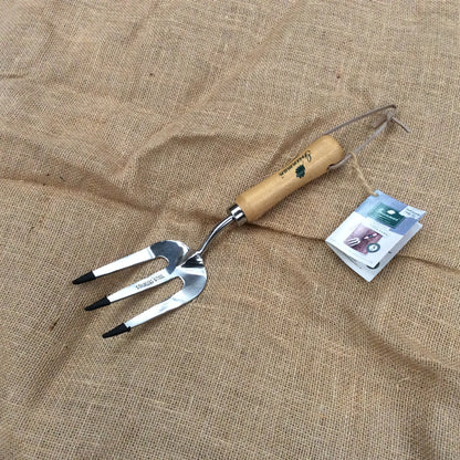 A metal garden hand fork with a light wood handle and black-tipped tines, placed on a burlap fabric background next to its product tag.